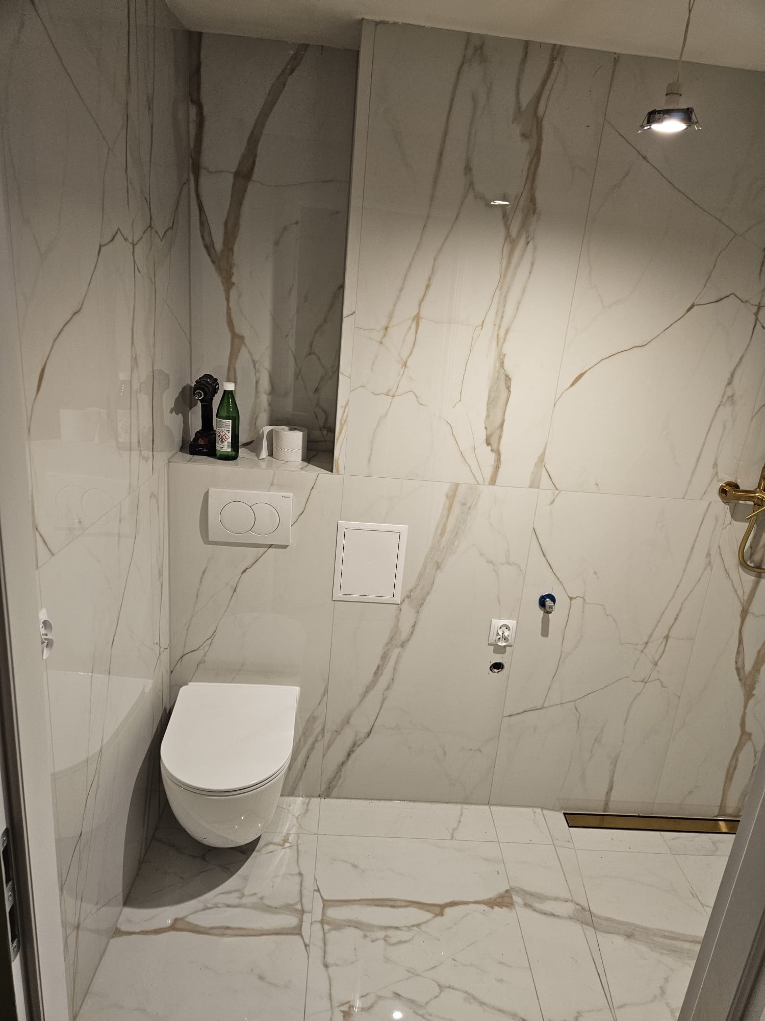 Nowoczesna toaleta w Tczewie z marmurowymi płytkami, podtynkową spłuczką i złotym odpływem liniowym. Widoczne narzędzia i akcesoria na półce.