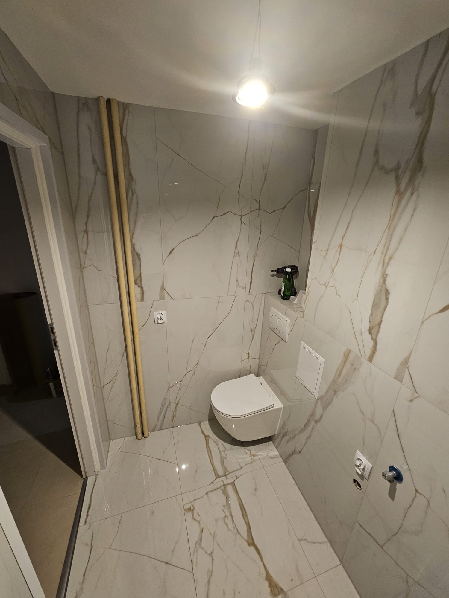 Wykończona łazienka z białymi płytkami imitującymi marmur, wiszącą toaletą i widocznymi rurami. W tle wiertarka na stelażu podtynkowym.