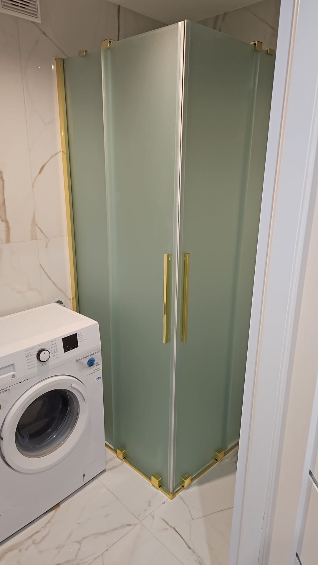 Szklana kabina prysznicowa z złotymi detalami w rogu łazienki, obok pralka. Marmurowe płytki podłogowe i ścienne.