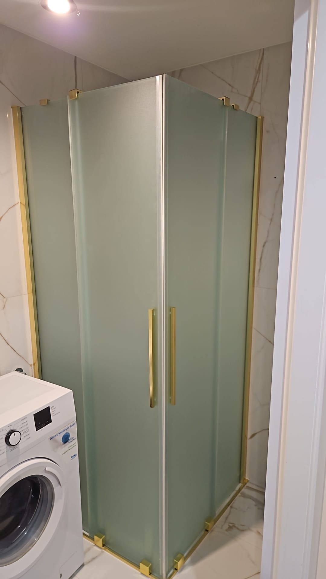 Nowoczesna kabina prysznicowa z matowego szkła i złotymi detalami w wykończonej łazience z marmurowymi płytkami i pralką. Minimalistyczny design.
