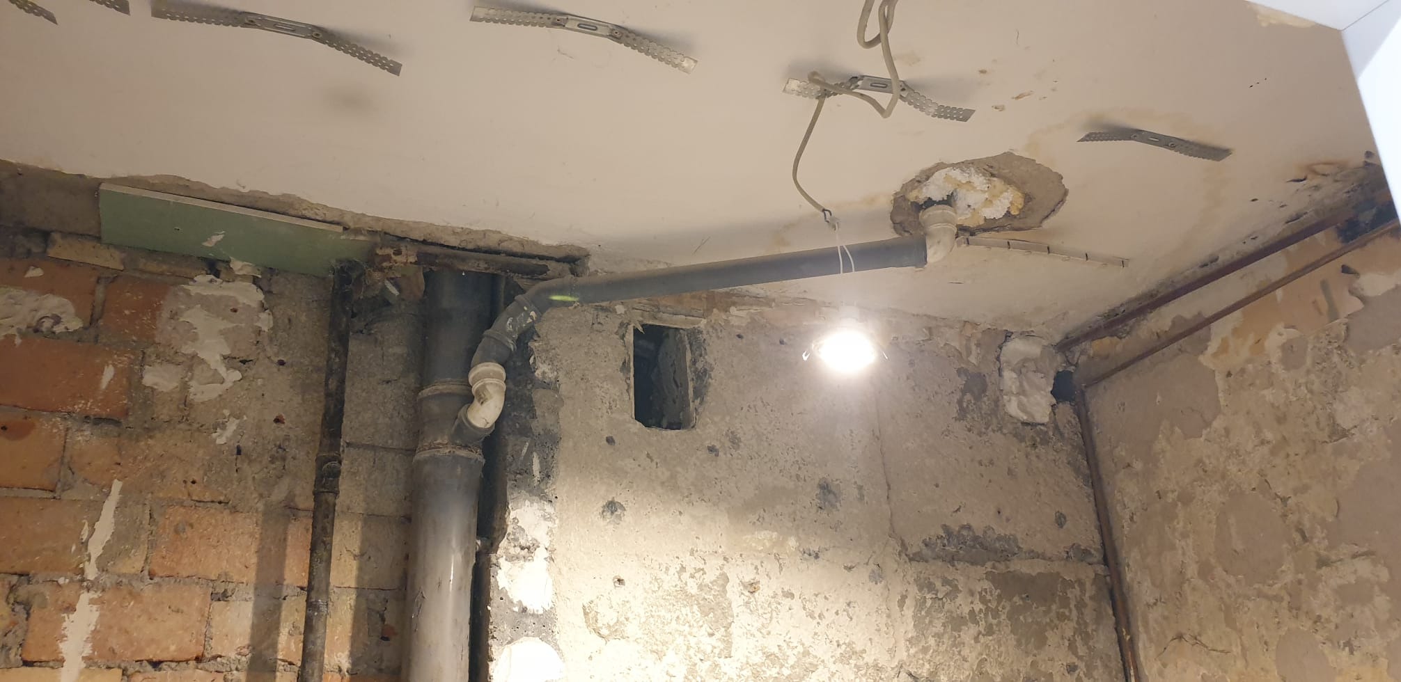 Fragment wnętrza w trakcie remontu: widoczne rury, instalacje pod sufitem, surowe ściany z ubytkami tynku i odsłoniętą cegłą, oświetlenie robocze.