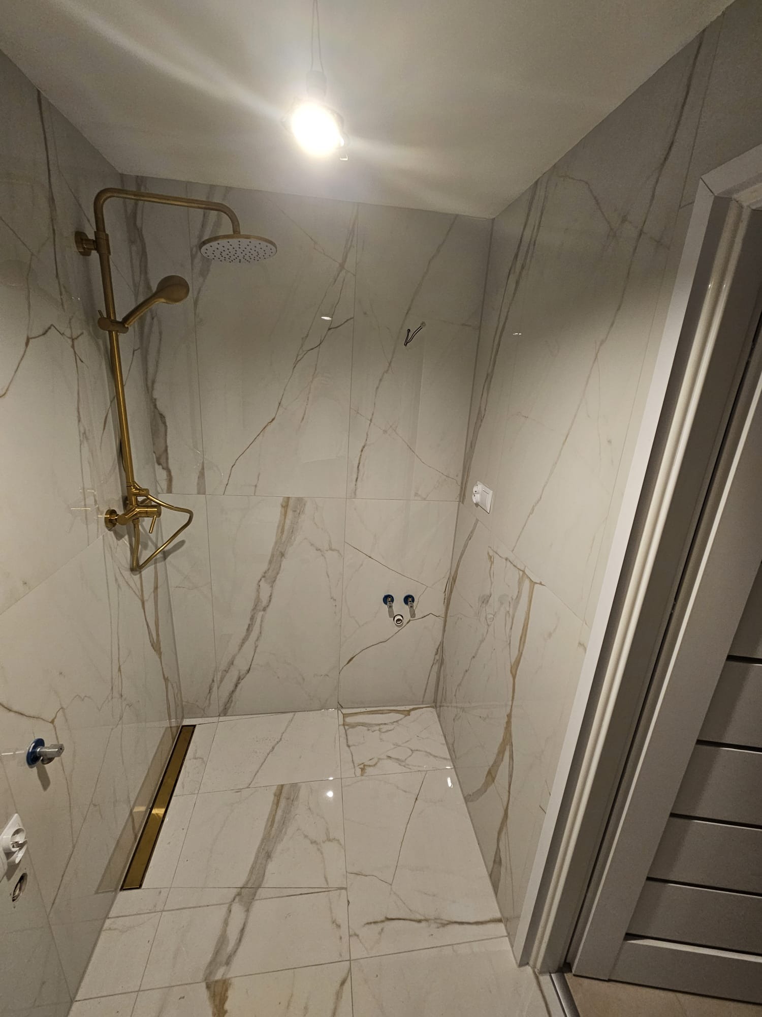 Wykończona kabina prysznicowa z marmurowymi płytkami, złotą armaturą i odpływem liniowym. Nowoczesny design, jasne oświetlenie, widok z góry.