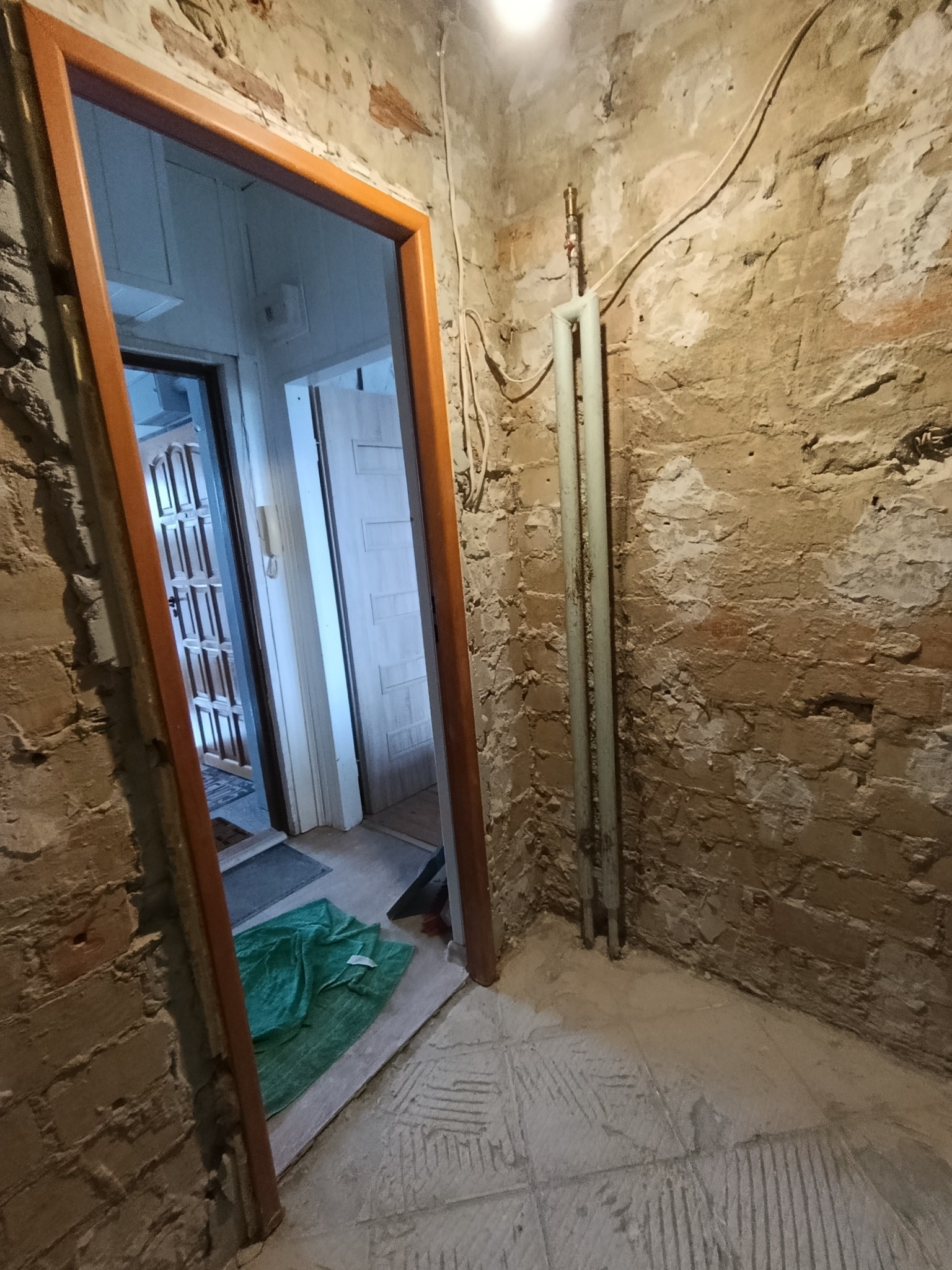 Wnętrze w trakcie remontu: odsłonięta cegła, grzejnik, otwarte drzwi z widokiem na dalsze pomieszczenia. Podłoga przygotowana do dalszych prac wykończeniowych.