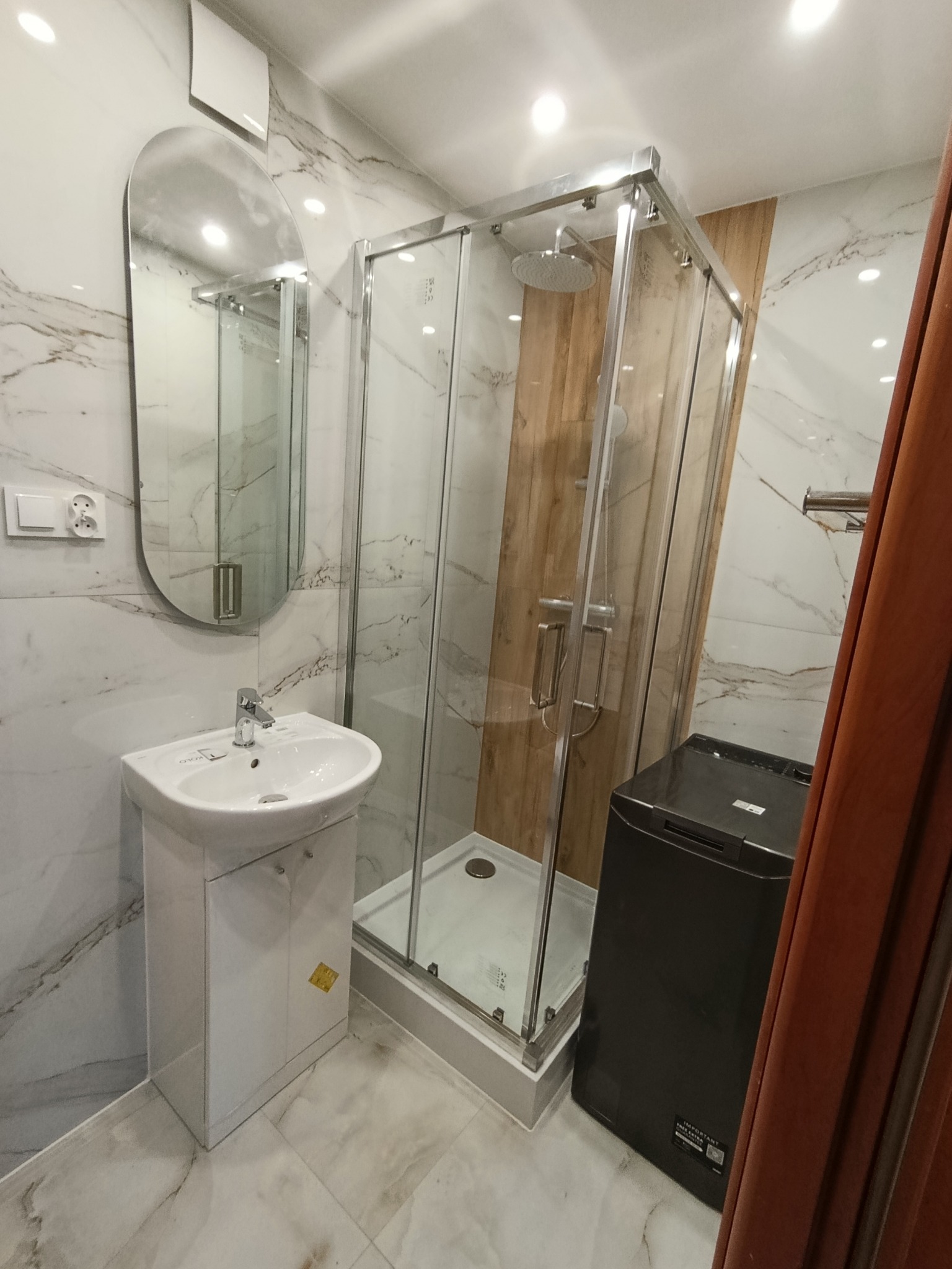 Nowoczesna łazienka z kabiną prysznicową, umywalką na szafce i pralką. Marmurowe płytki na ścianach i podłodze. Owalne lustro nad umywalką.