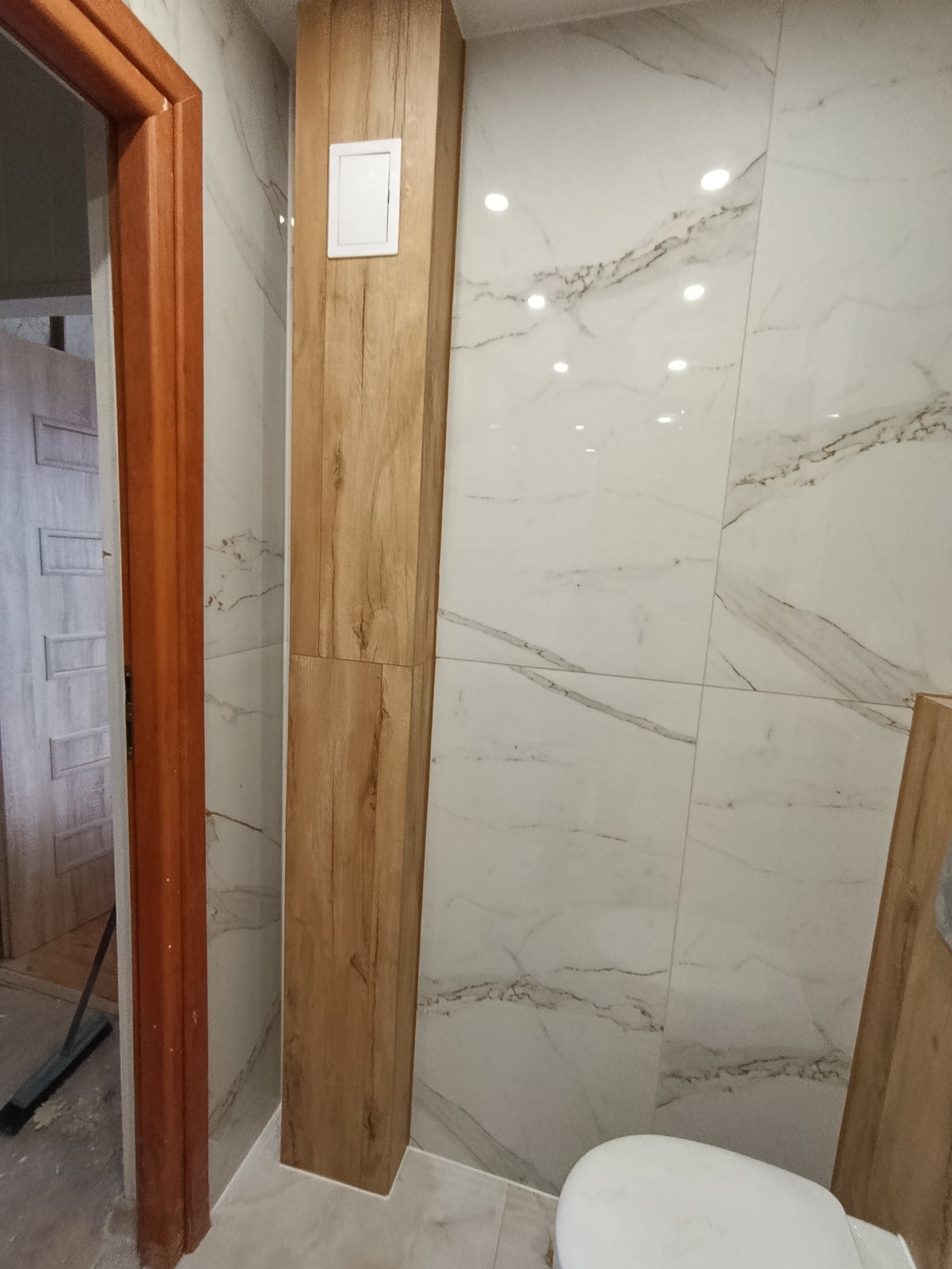 Wykończona łazienka z płytkami imitującymi marmur i drewnopodobną obudową pionu z włącznikiem. Widoczny fragment toalety i ościeżnica drzwi.