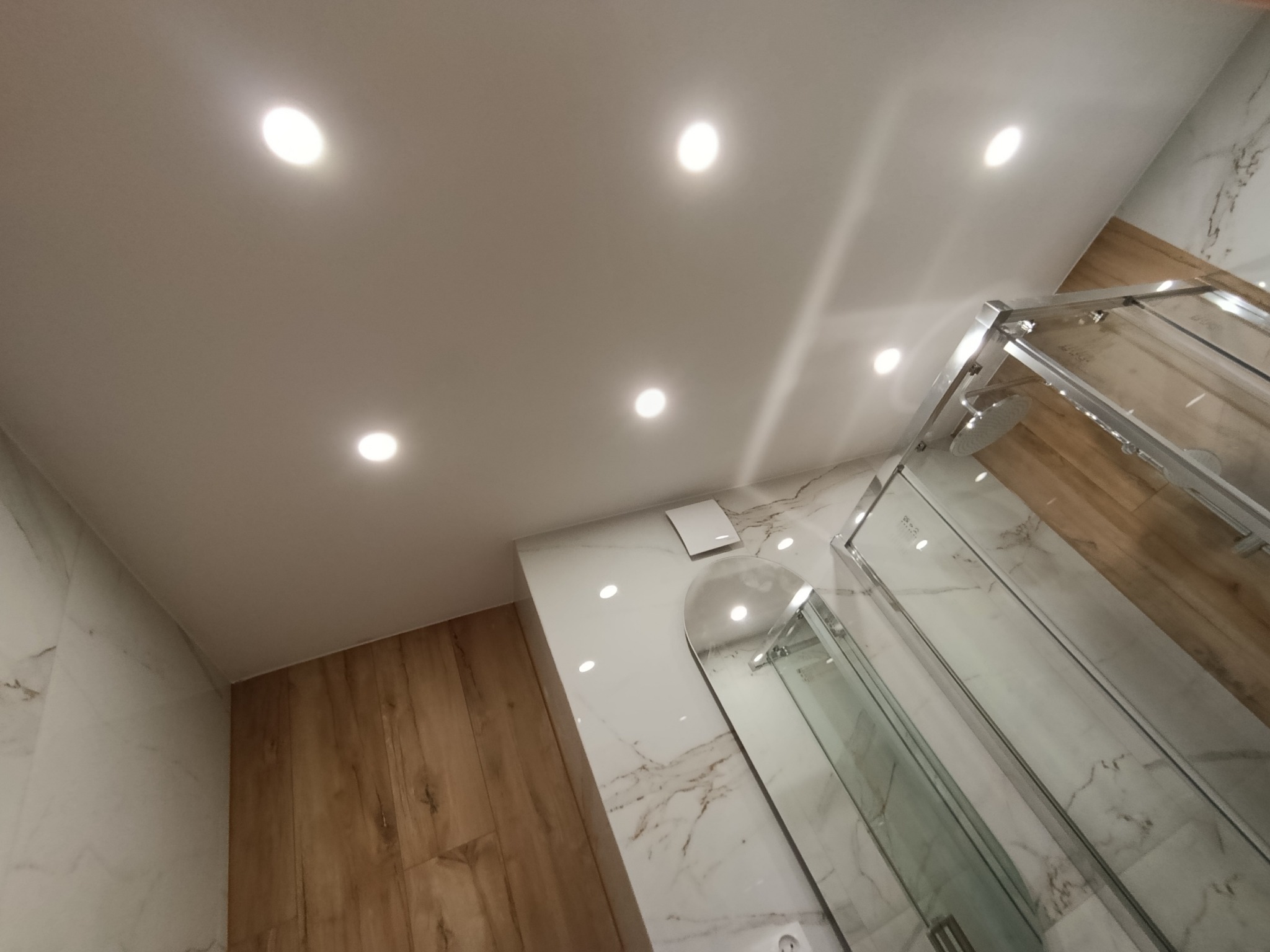 Nowoczesna kabina prysznicowa z białymi płytkami imitującymi marmur i drewnopodobnymi panelami w Tczew. Sufit z oświetleniem punktowym.