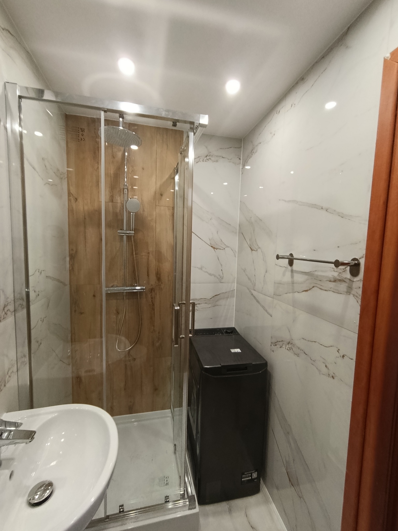 Wykończona łazienka z kabiną prysznicową typu walk-in z drewnianym panelem, umywalką i pralką. Marmurowe płytki na ścianach i podłodze. Nowoczesny design.