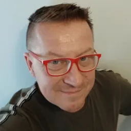 Portret mężczyzny w czerwonych okularach, z krótkimi włosami i lekkim uśmiechem, skierowany w stronę obiektywu na jasnym tle.