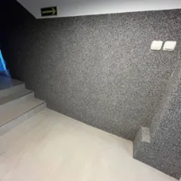 Ściana z szarego mikrocementu w klatce schodowej, z widocznymi schodami i zielonym znakiem ewakuacyjnym. Dwa włączniki światła na ścianie.