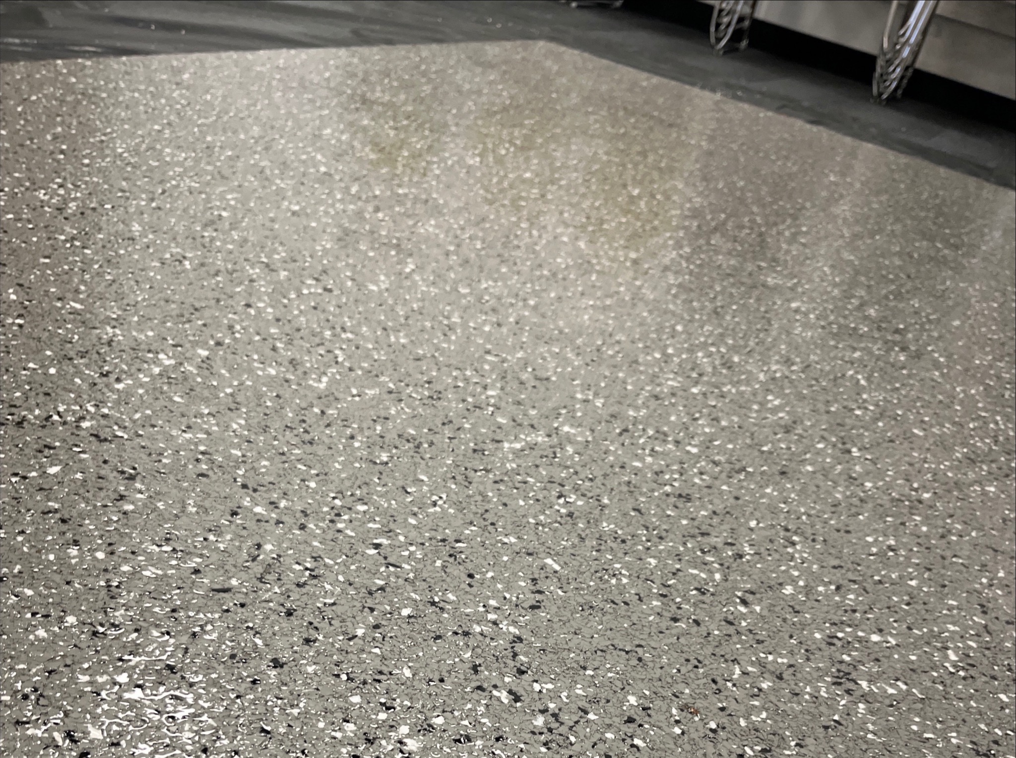 Błyszcząca, szara posadzka żywiczna z czarno-białymi drobinkami, widoczna w perspektywie z bliska, z fragmentem metalowej konstrukcji w tle.