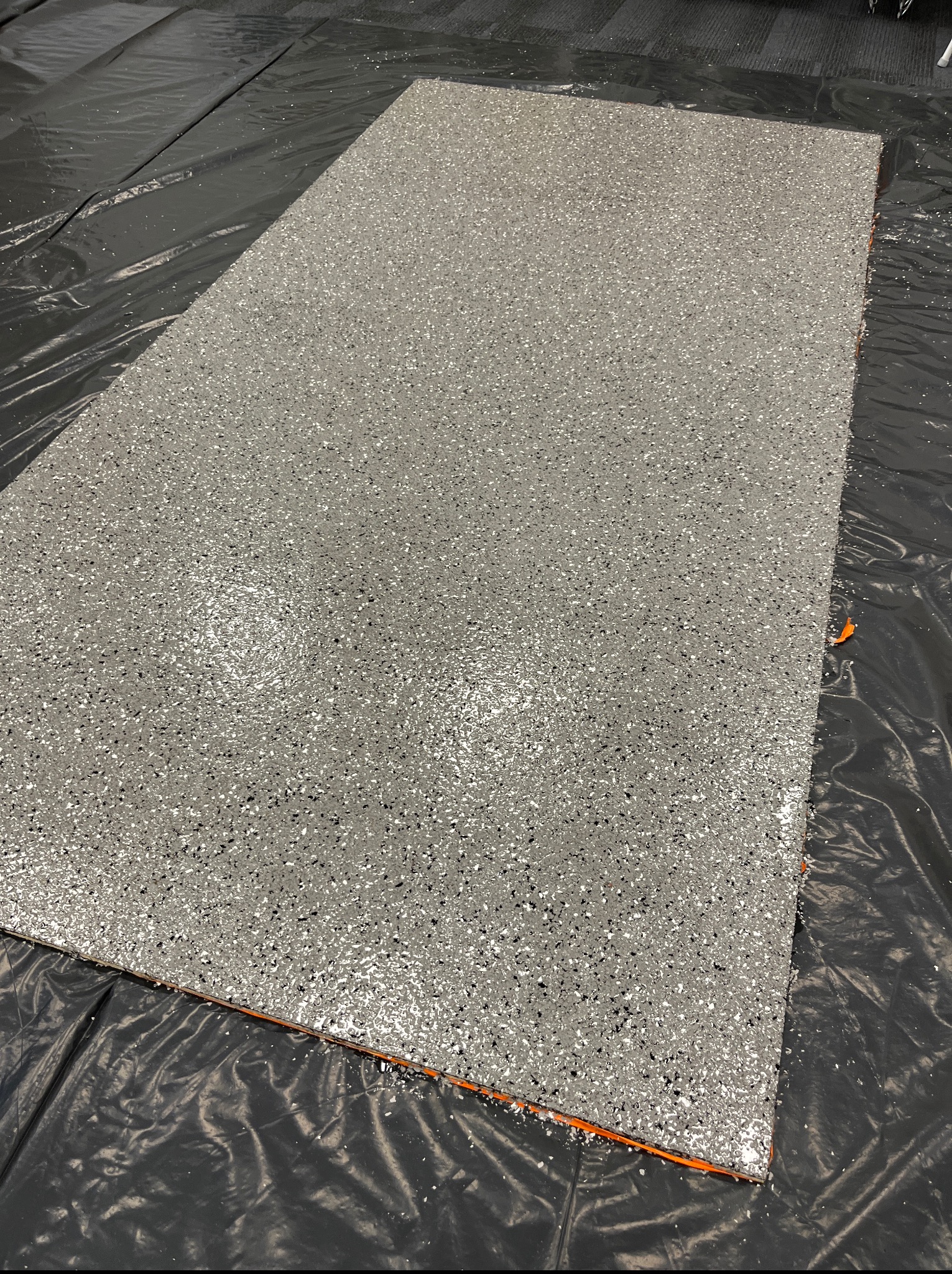 Świeżo wylana, szara posadzka żywiczna z brokatem, zabezpieczona folią ochronną. Widoczne drobne nierówności i refleksy świetlne na powierzchni.