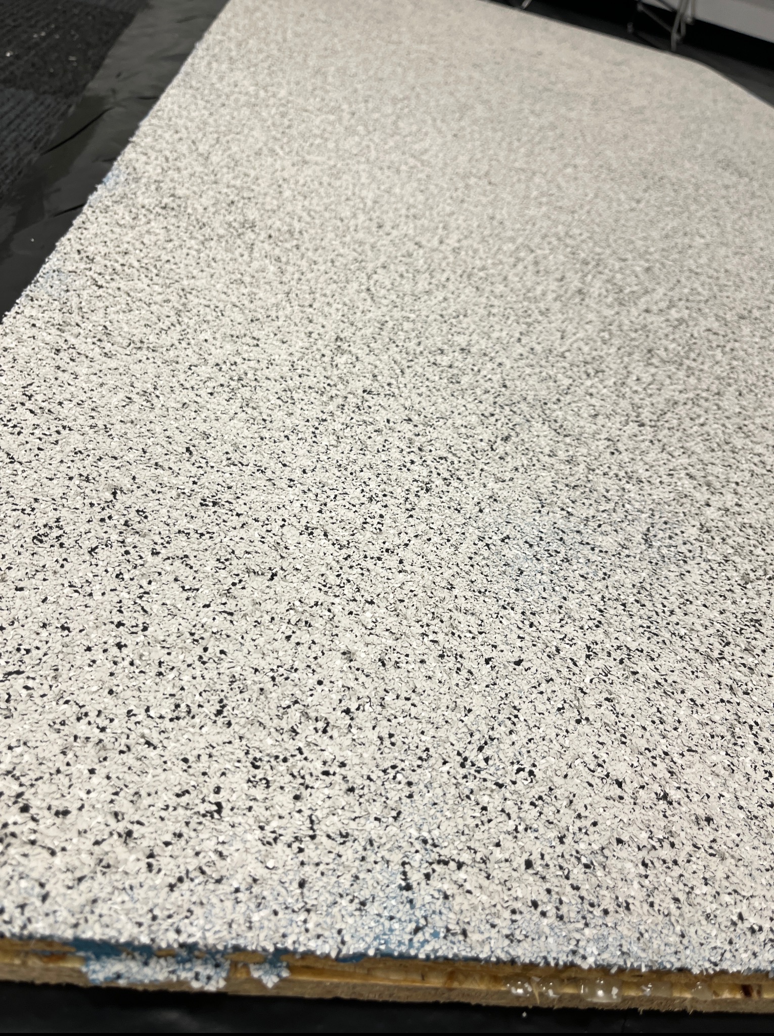 Jasnoszara posadzka żywiczna z drobnymi, ciemnymi elementami, ukazana pod kątem, z widoczną krawędzią podłoża. Powierzchnia matowa, bez refleksów.