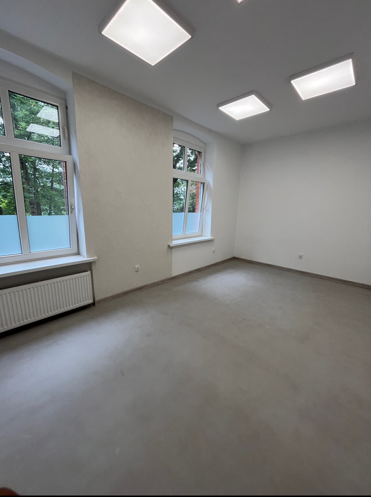 Jasne wnętrze po remoncie: gładkie, szare posadzki, białe ściany z delikatną fakturą, okna z widokiem na zieleń. Minimalistyczny design, równomierne oświetlenie.