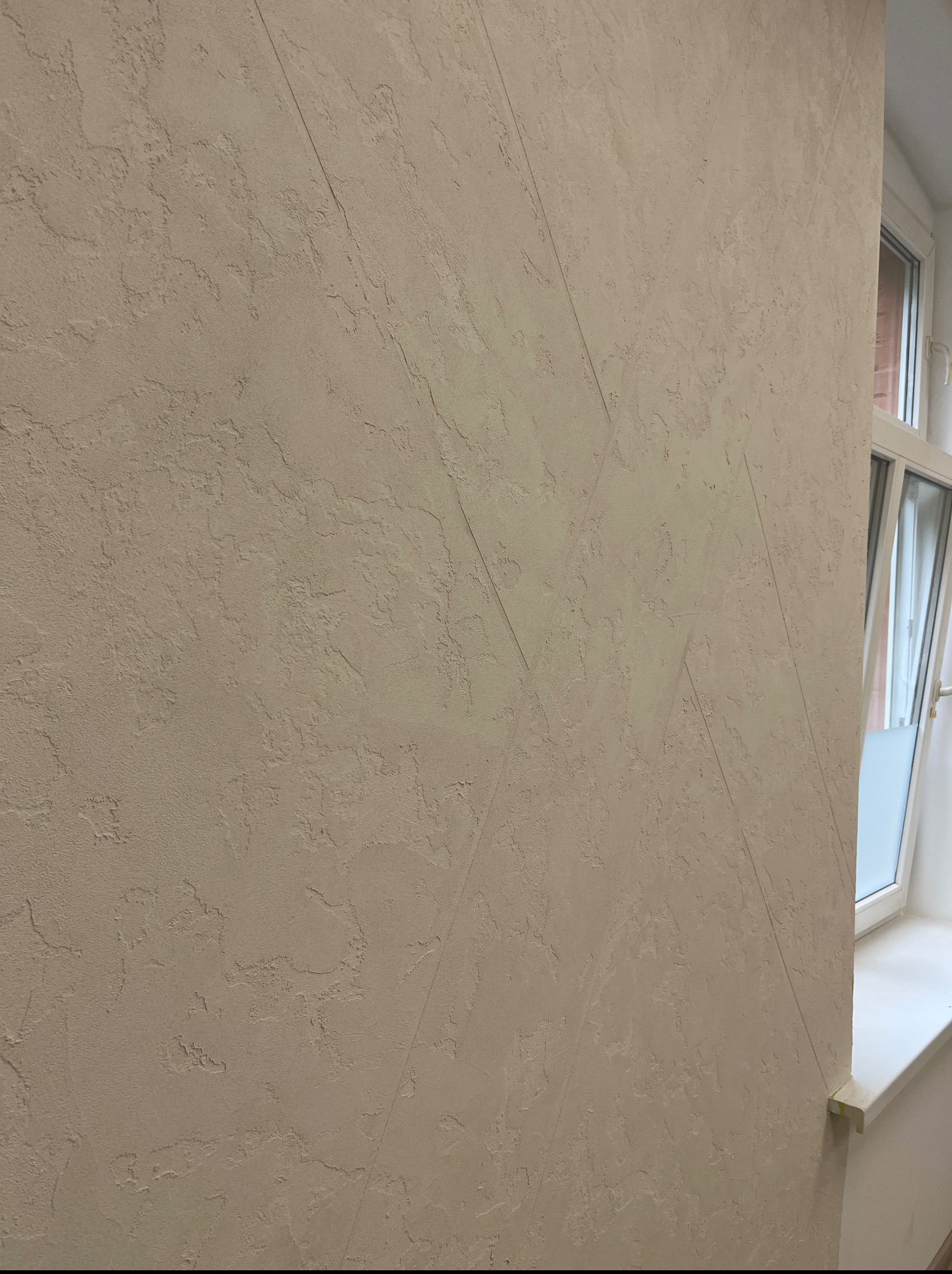 Beżowa ściana z dekoracyjną fakturą i delikatnymi, ukośnymi liniami, przylegająca do białego okna. Tekstura ściany jest nierówna, z subtelnymi wgłębieniami i wypukłościami.