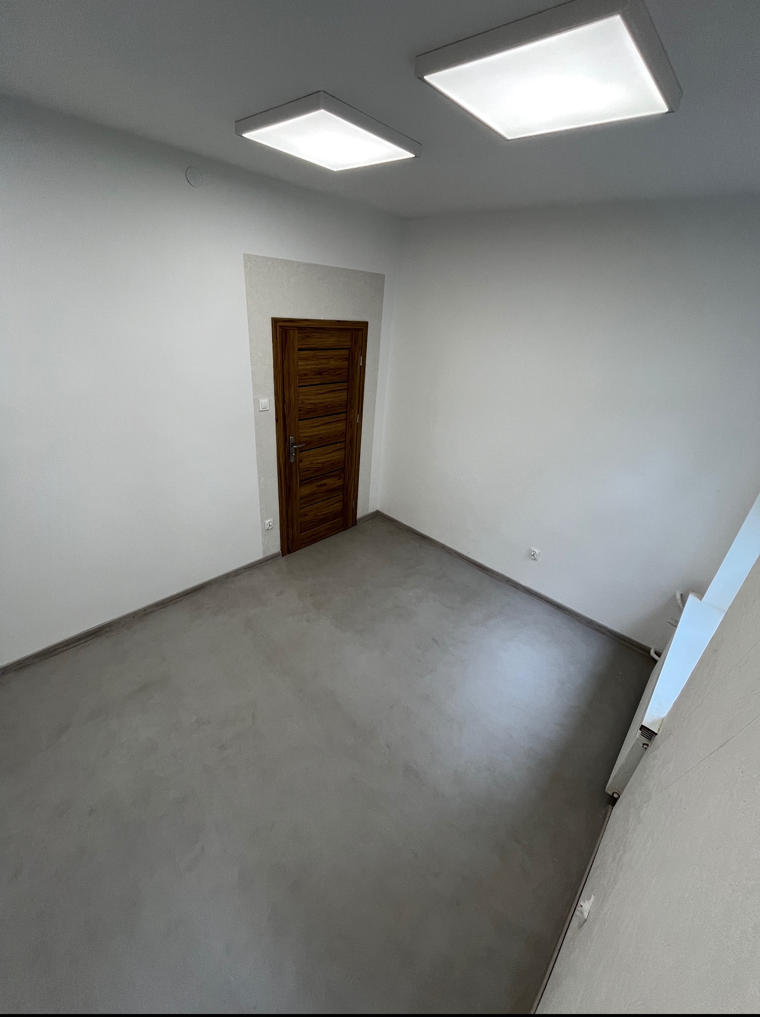 Szara posadzka mikrocementowa w pustym, jasnym pomieszczeniu z drewnianymi drzwiami i dwoma kwadratowymi lampami sufitowymi. Widok z góry.