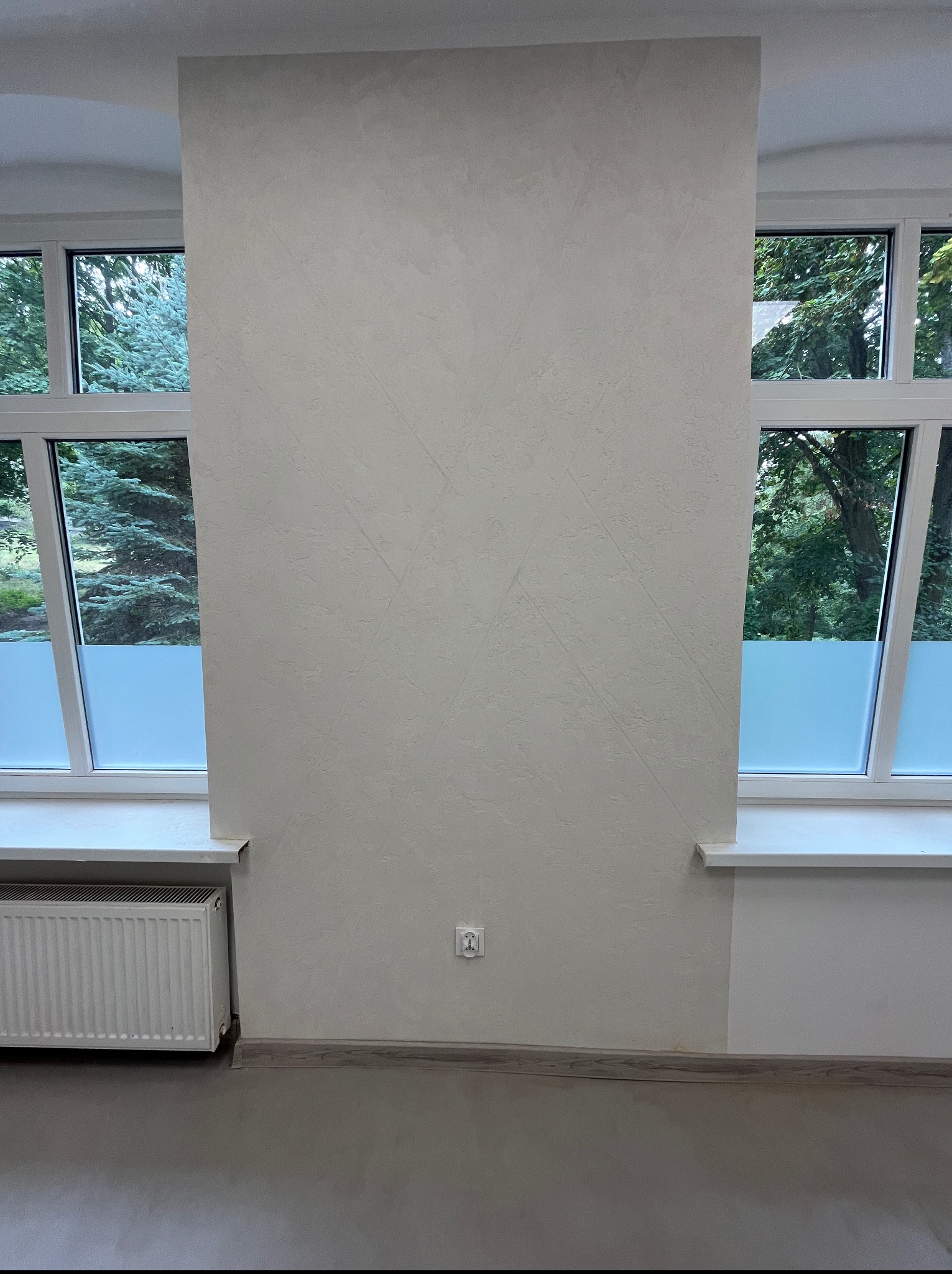 Ściana z dekoracyjną, jasną fakturą, widoczna między dwoma oknami z widokiem na zieleń. Grzejnik i listwa przypodłogowa uzupełniają wnętrze.