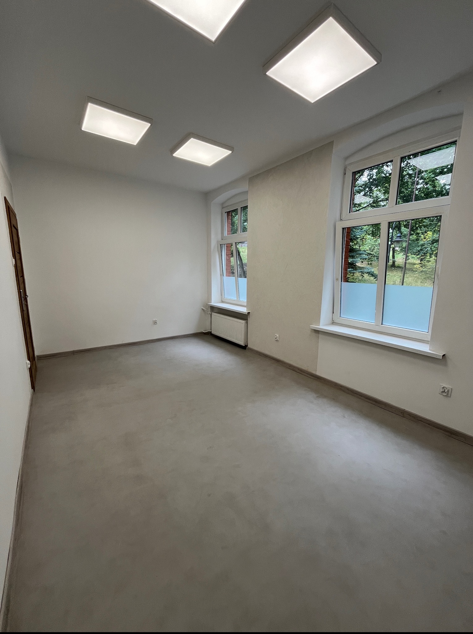 Wnętrze pokoju z szarą posadzką, trzema oknami z częściowo zamontowaną folią i kwadratowymi lampami sufitowymi. Widok na zewnątrz przez okna.