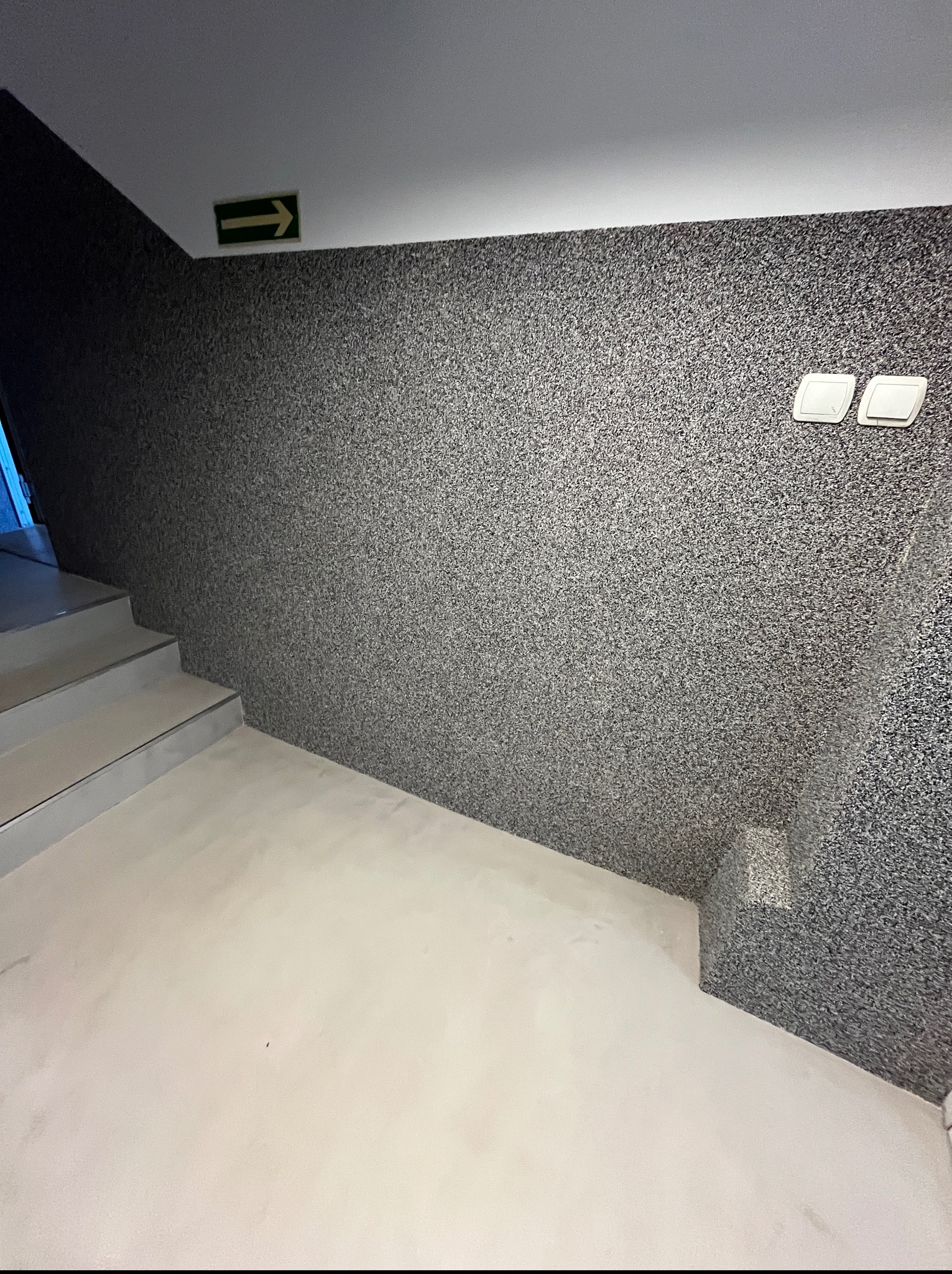 Ściana z szarego mikrocementu w klatce schodowej, z widocznymi schodami i zielonym znakiem ewakuacyjnym. Dwa włączniki światła na ścianie.