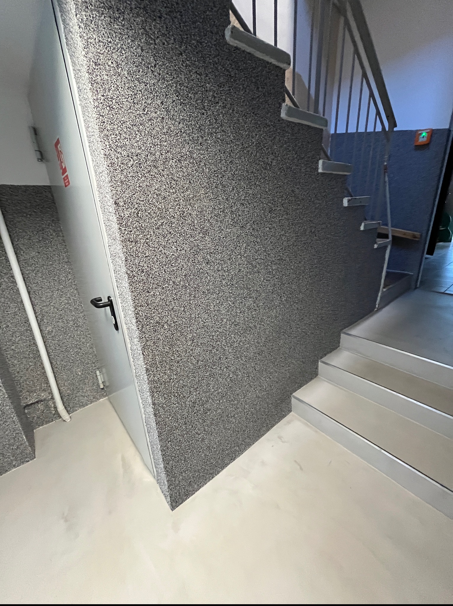 Klatka schodowa w budynku użyteczności publicznej. Ściany i schody pokryte szarą, dekoracyjną posadzką żywiczną. Na ścianie widoczne drzwi przeciwpożarowe.