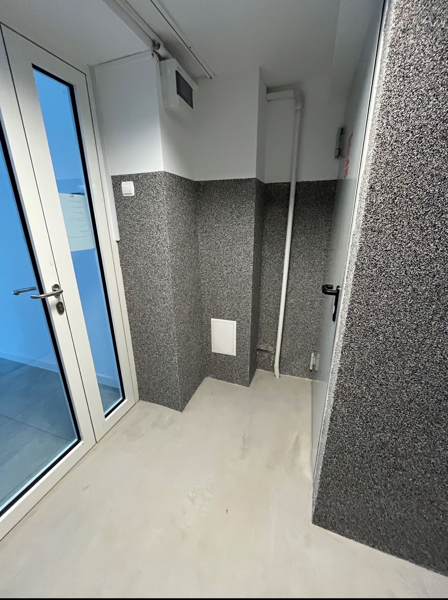 Wnętrze po remoncie: szara, teksturowana ściana do połowy wysokości, jasna podłoga i białe drzwi z widokiem na zewnątrz. Minimalistyczny design.