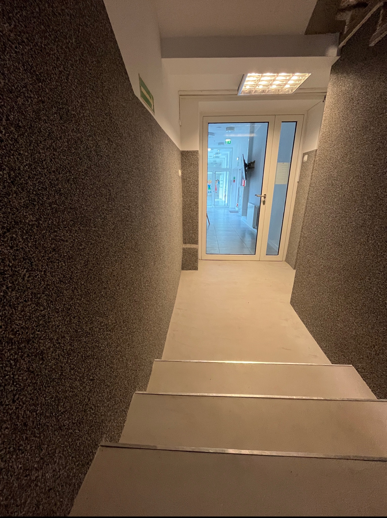 Klatka schodowa wykończona szpachlą dekoracyjną w odcieniach brązu, prowadząca do przeszklonych drzwi. Widoczny fragment schodów i oświetlenie sufitowe.