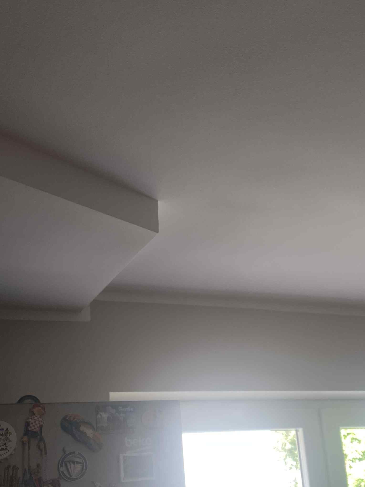 Gładki, biały sufit z subtelnym oświetleniem i dekoracyjnym elementem architektonicznym. Widoczny fragment ściany i okna w tle.