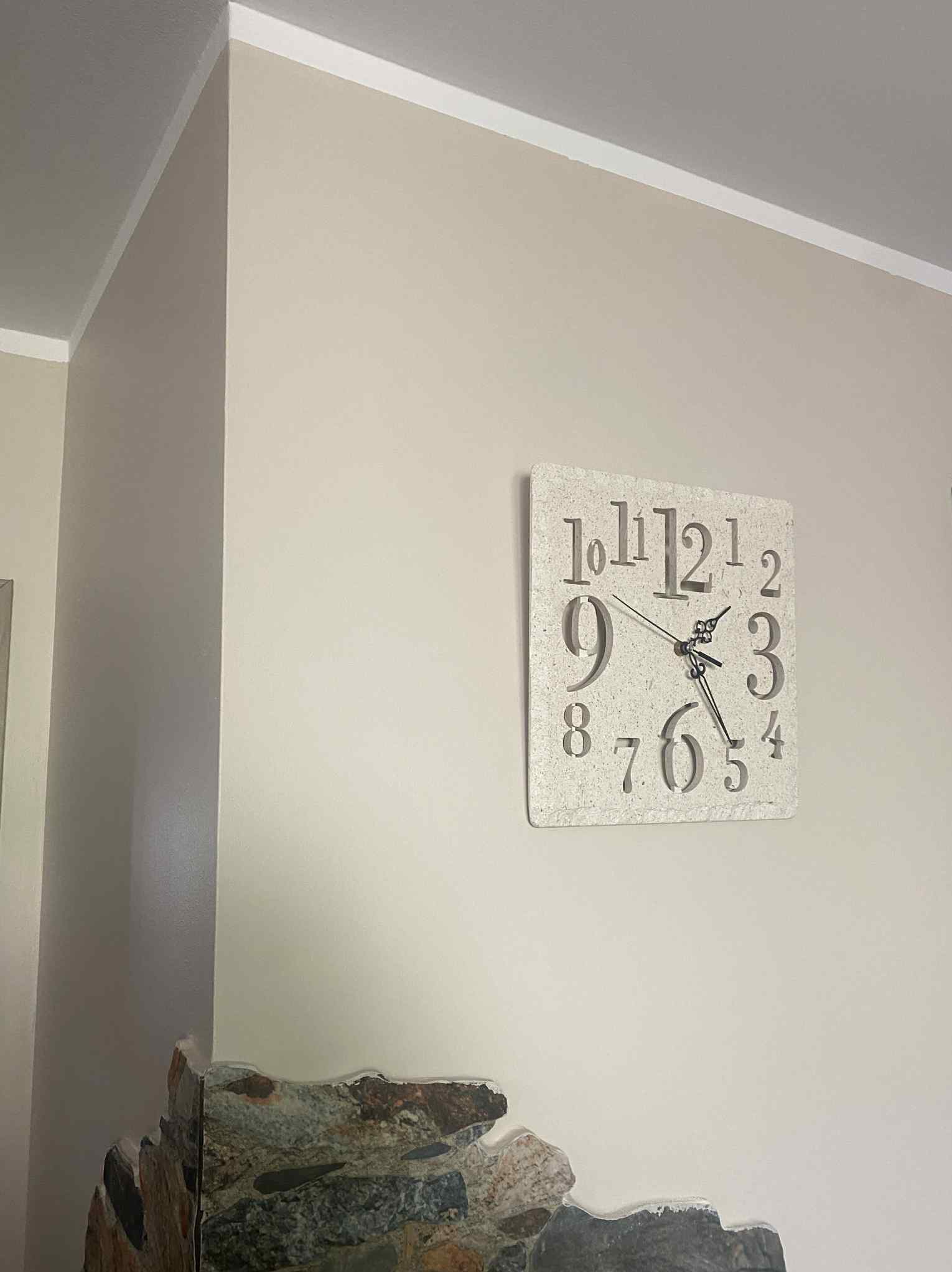 Jasna ściana po malowaniu z dekoracyjnym zegarem. Widoczny fragment kamiennej dekoracji ściany. Neutralna kolorystyka wnętrza, wykończenie listwą przysufitową.