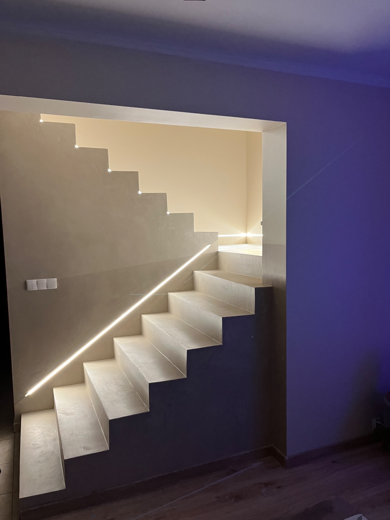 Nowoczesne schody z mikrocementu z subtelnym oświetleniem LED, kremowe ściany i drewniana podłoga. Minimalistyczny design z akcentem na detale wykończeniowe.