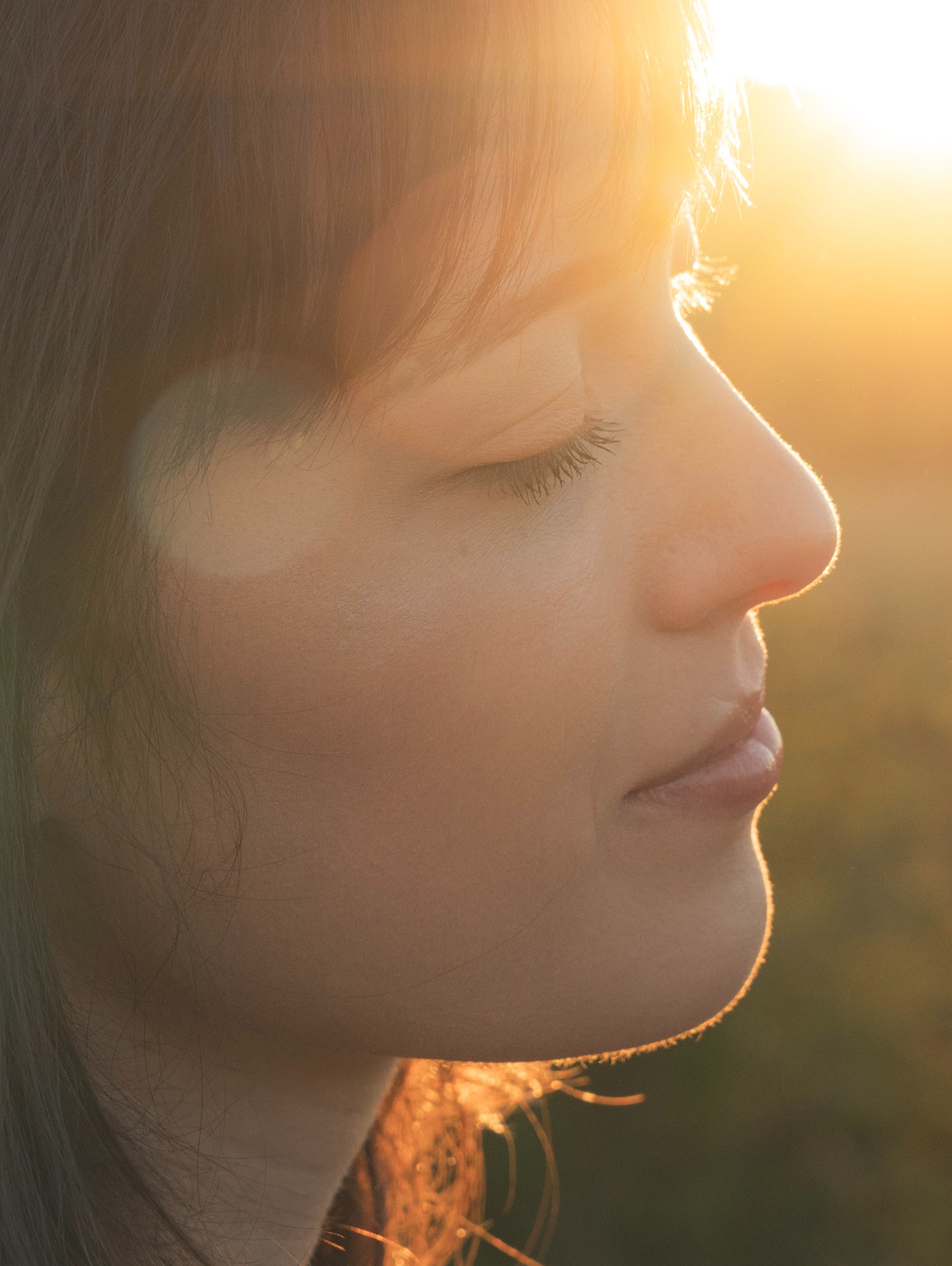 Profil kobiety z zamkniętymi oczami, oświetlony ciepłym, złotym światłem słonecznym, tworzącym subtelny blask na skórze i włosach.