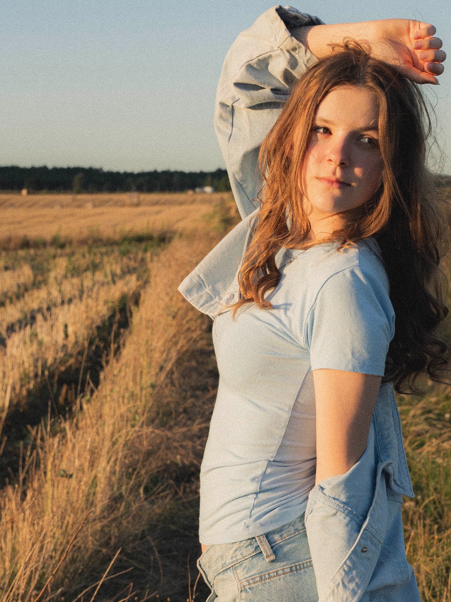 Portret młodej kobiety w dżinsowej kurtce na tle pola w ciepłym, zachodzącym słońcu. Naturalny makijaż, swobodna poza i miękkie światło.