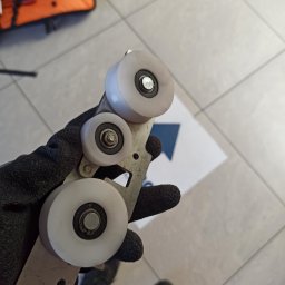 ACCUGATE SERWIS SZYMON MARCZAK - Ręka w czarnej rękawicy trzyma metalowy element z trzema białymi rolkami, prawdopodobnie część mechanizmu bramy przesuwnej. Widoczne ślady użytkowania.