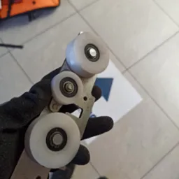 Ręka w czarnej rękawicy trzyma metalowy element z trzema białymi rolkami, prawdopodobnie część mechanizmu bramy przesuwnej. Widoczne ślady użytkowania.