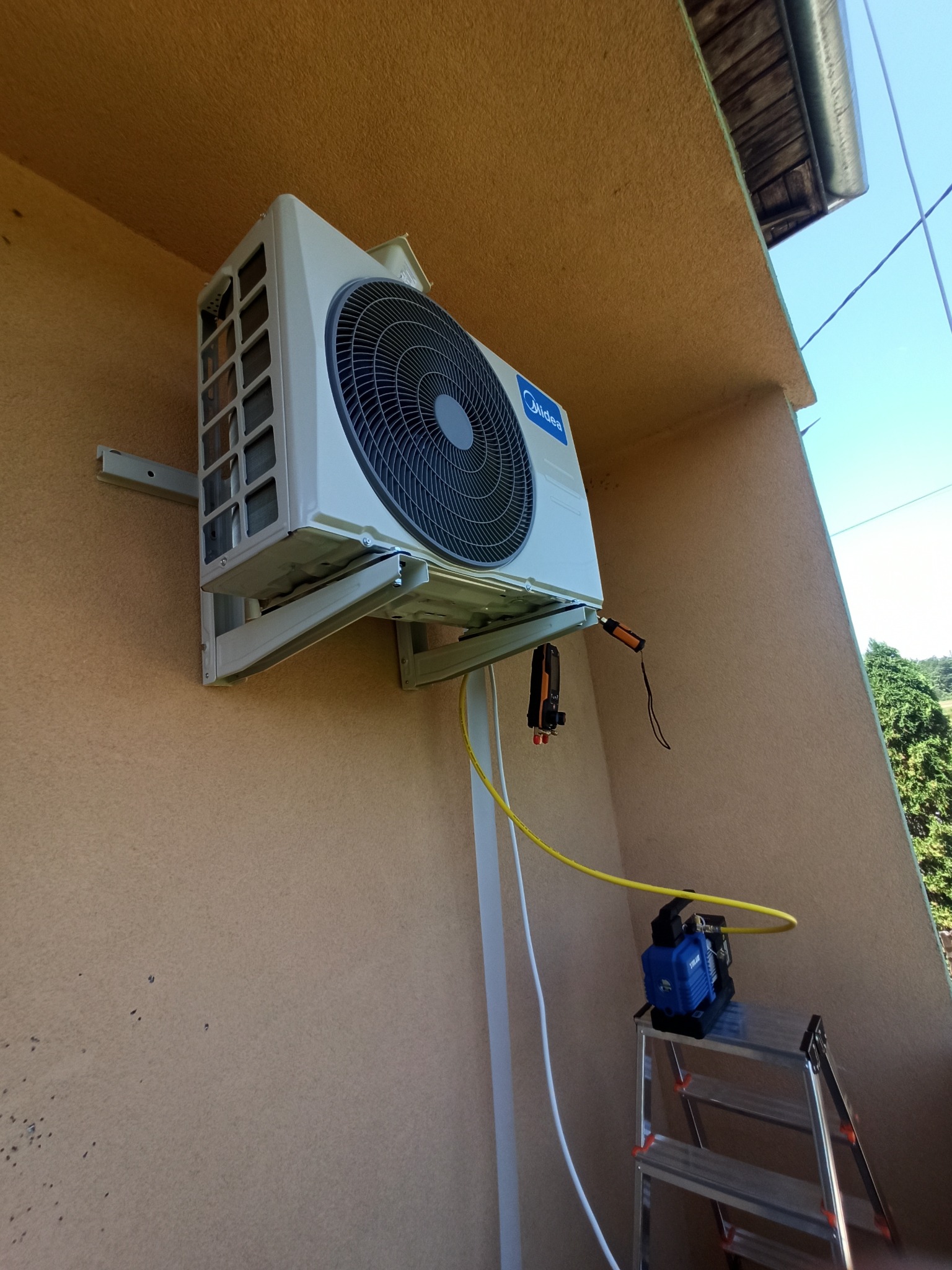 Biały klimatyzator Midea zamontowany na ścianie budynku, widoczny podczas serwisu z użyciem pompy próżniowej i drabiny. Żółty przewód łączy urządzenie z klimatyzatorem.