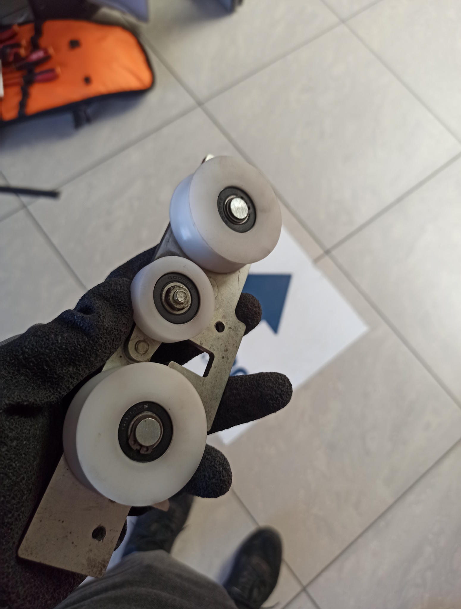 Ręka w czarnej rękawicy trzyma metalowy element z trzema białymi rolkami, prawdopodobnie część mechanizmu bramy przesuwnej. Widoczne ślady użytkowania.