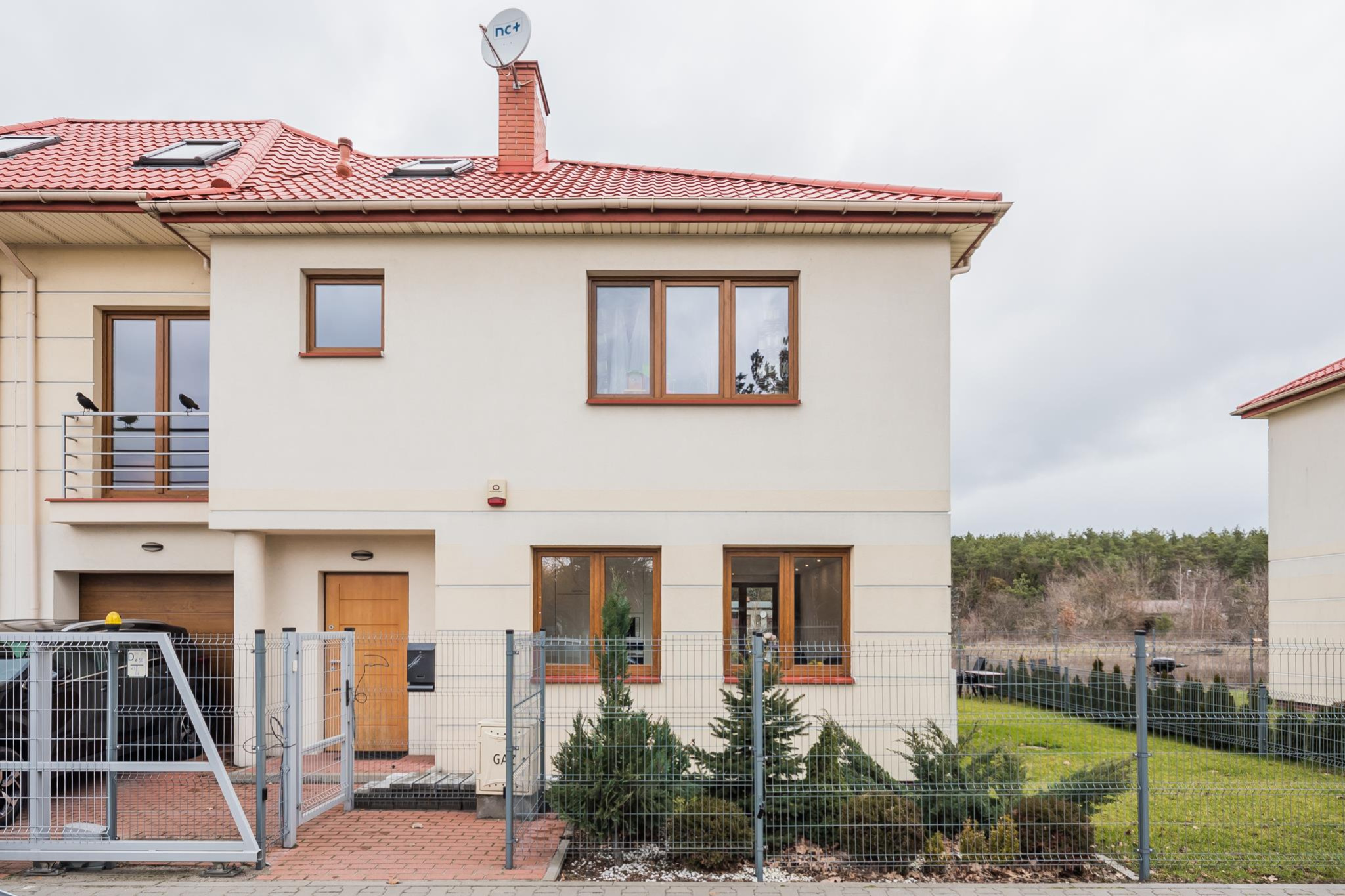 Dwukondygnacyjny dom szeregowy z czerwonym dachem i brązowymi oknami. Na balkonie siedzą dwa kruki. Widoczny fragment ogrodzenia i zieleni.