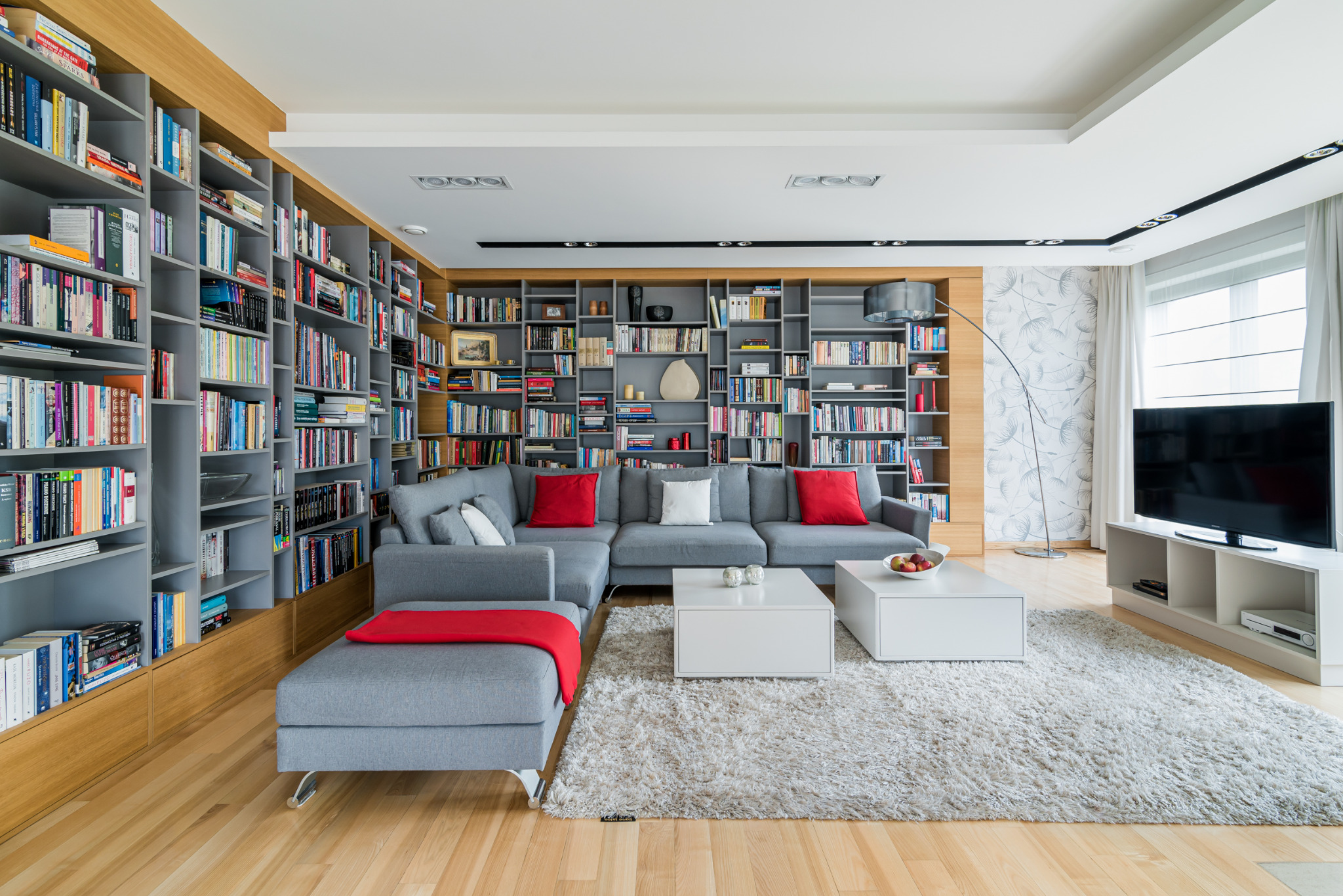 Przestronny salon z szarym narożnikiem, białymi stolikami i regałami pełnymi książek. Czerwone akcenty na poduszkach i pledzie dodają energii. Jasne drewno podłogi.