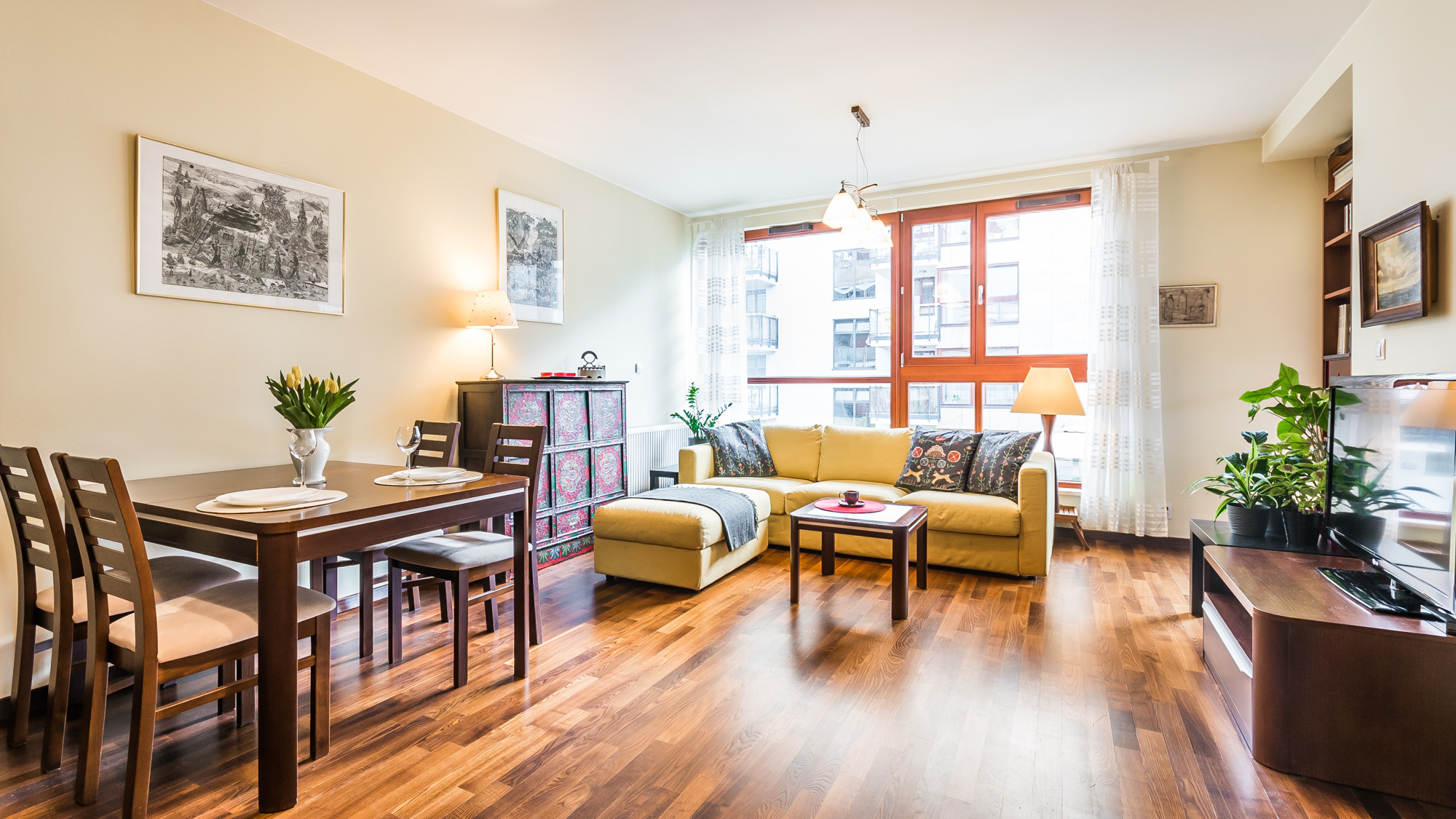 Jasne wnętrze mieszkania z drewnianą podłogą, żółtą sofą, stołem z krzesłami i dużym oknem. Na stole wazon z tulipanami. Przy oknie widok na inny budynek.