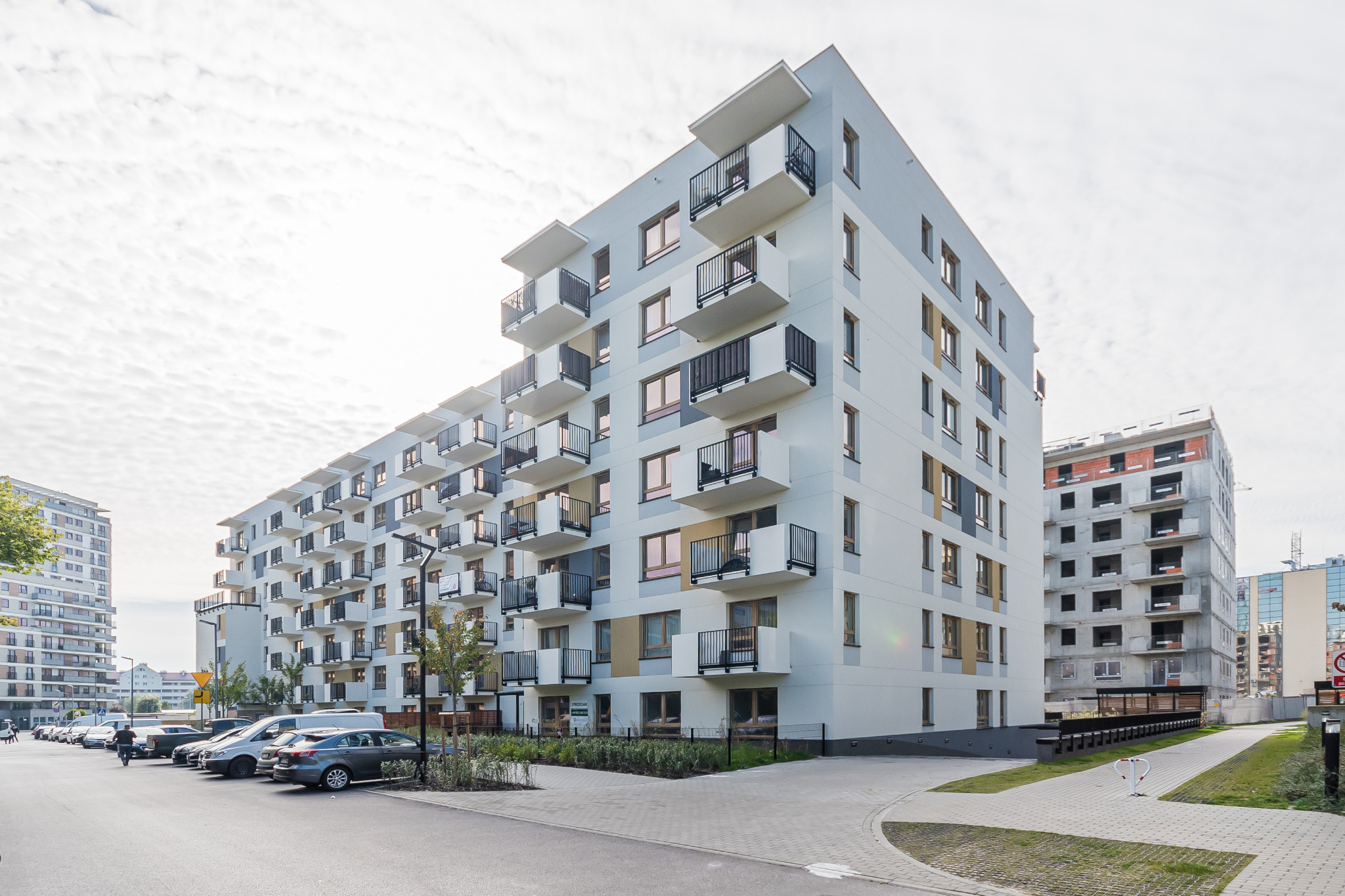 Nowoczesny, jasny apartamentowiec z balkonami i zaparkowanymi autami wzdłuż chodnika. W tle budynek w stanie surowym.