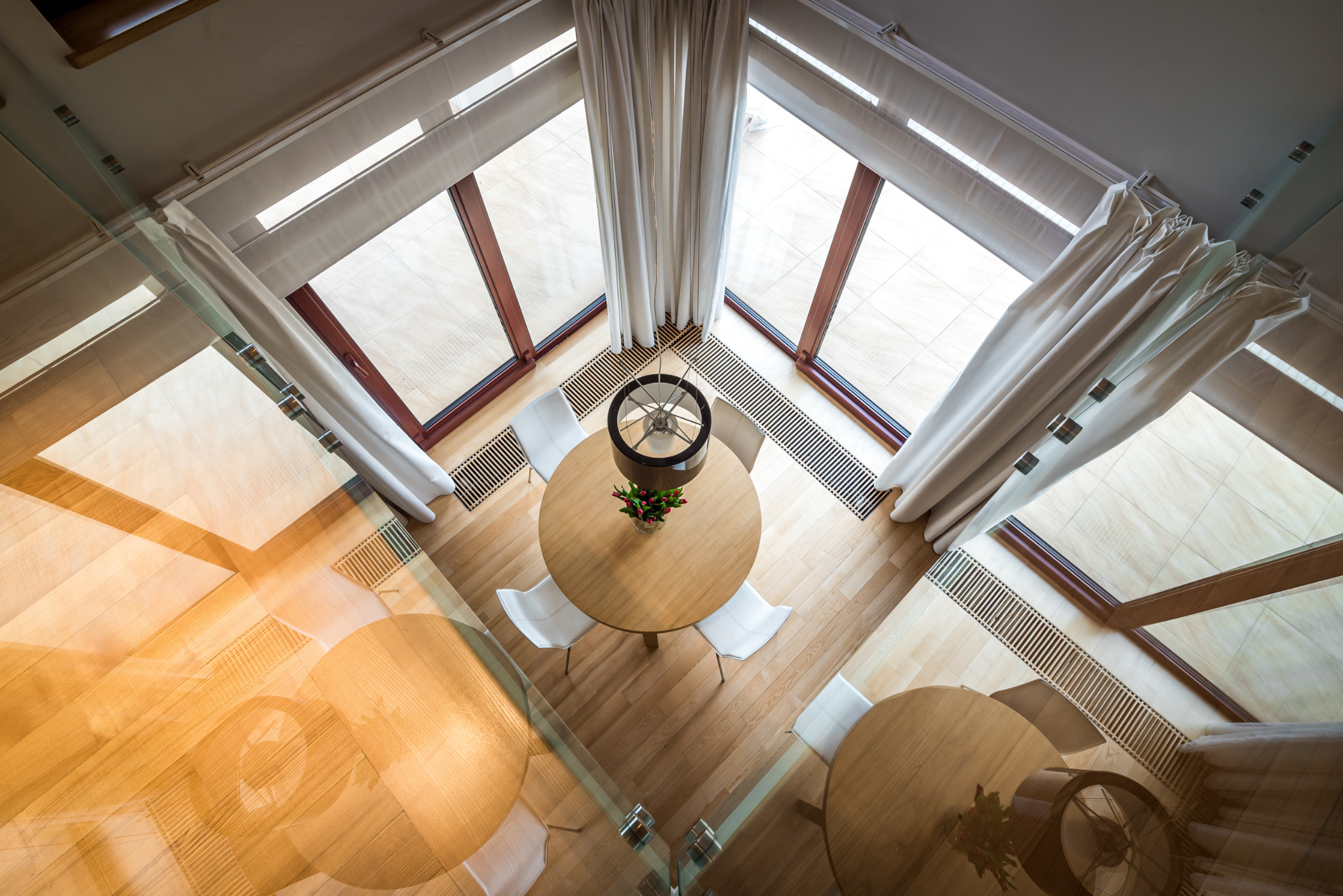 Nowoczesne wnętrze z drewnianą podłogą, okrągłym stołem i białymi krzesłami, widziane z góry przez szklaną balustradę. Duże okna z jasnymi zasłonami.