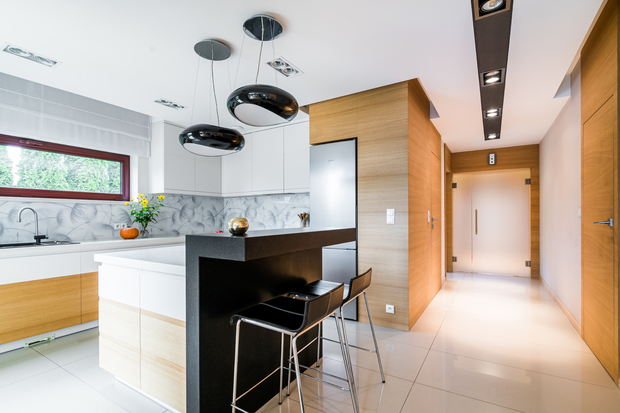 Nowoczesna kuchnia z wyspą i hokerami, białe szafki z drewnianymi akcentami, czarny blat, designerskie lampy wiszące, widok na korytarz z drewnianym wykończeniem.