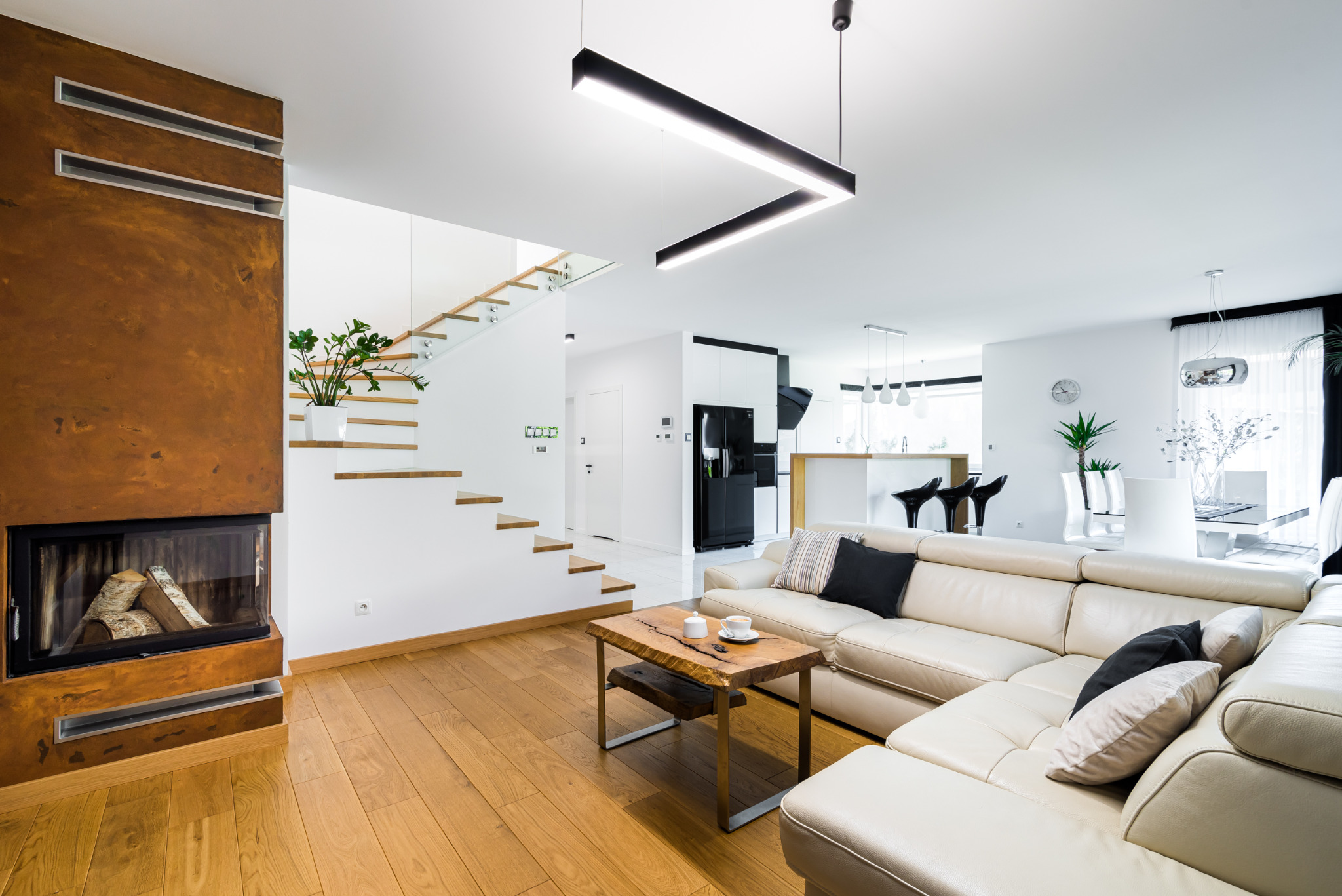 Jasne, przestronne wnętrze domu z kominkiem o rdzawym wykończeniu, drewnianymi schodami i narożną sofą. Nowoczesne oświetlenie i minimalistyczny design.