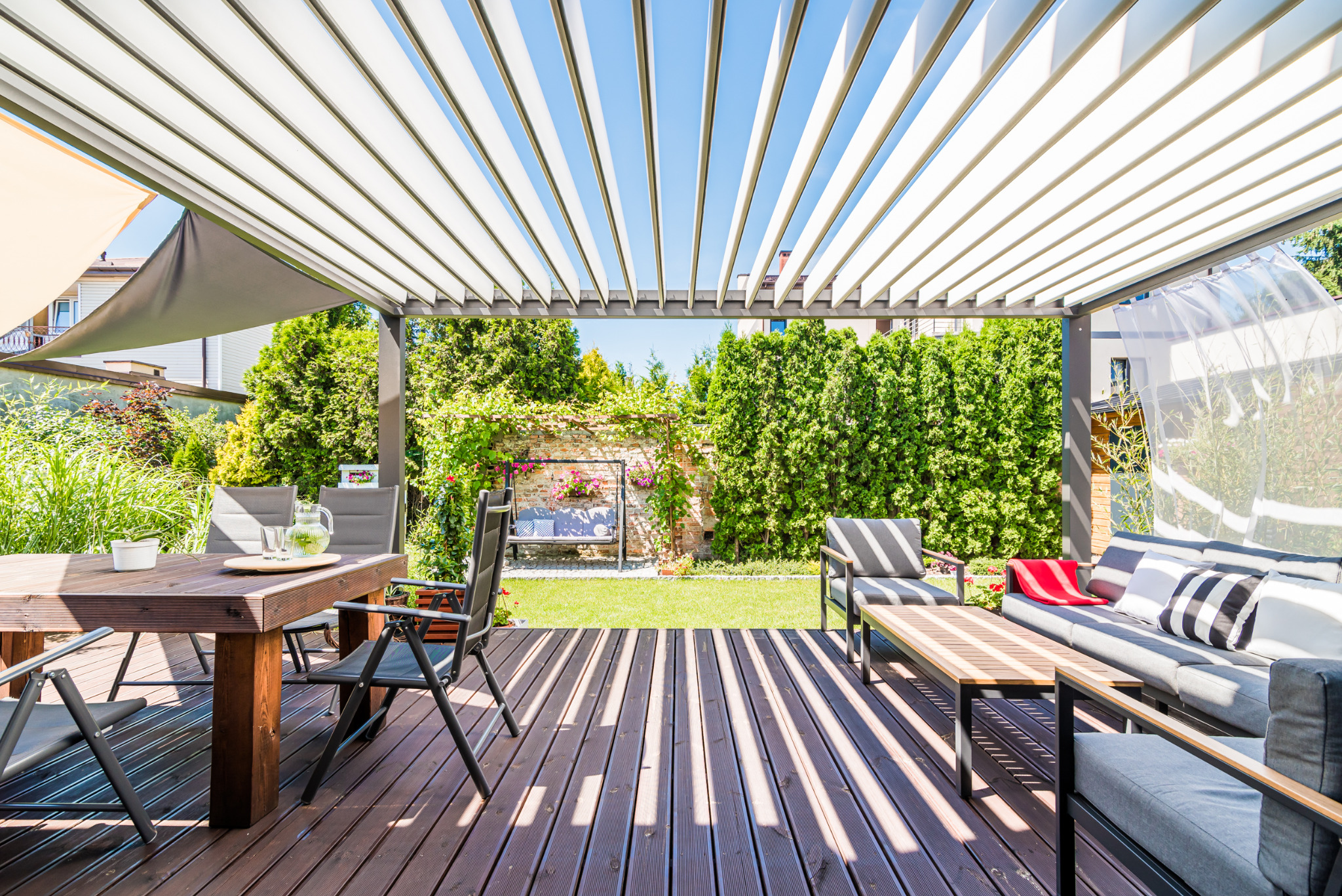 Zadaszony taras z drewnianą podłogą, meblami ogrodowymi i widokiem na zielony ogród. Pergola z białymi lamelami tworzy cień. Idealne miejsce do relaksu na świeżym powietrzu.