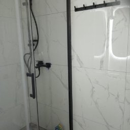 Kamil libner - Wykończona kabina prysznicowa z czarną armaturą i uchwytem na ręczniki na tle białych płytek imitujących marmur. Widoczne akcesoria i wiadro po remoncie.