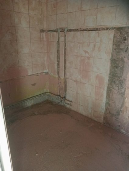 Łazienka w trakcie remontu: stare płytki częściowo usunięte, rury na ścianie, wylewka na podłodze. Widoczne ślady wcześniejszych prac i przygotowanie do dalszych etapów.