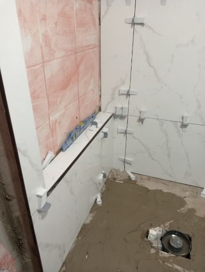 Białe płytki imitujące marmur układane z użyciem krzyżyków dystansowych, poziomica na krawędzi. Etap prac wykończeniowych w łazience.