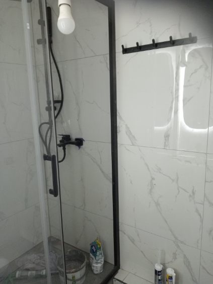 Wykończona kabina prysznicowa z czarną armaturą i uchwytem na ręczniki na tle białych płytek imitujących marmur. Widoczne akcesoria i wiadro po remoncie.