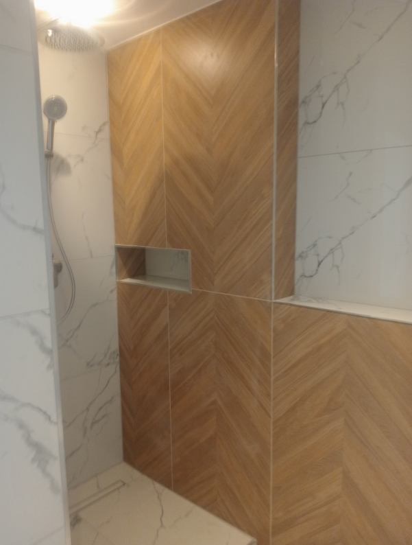 Nowoczesna kabina prysznicowa z drewnopodobnymi płytkami ułożonymi w jodełkę i marmurowymi ścianami. W ścianie wnęka na kosmetyki. Deszczownica i słuchawka prysznicowa.