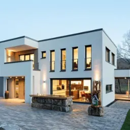 Nowoczesny dom z białymi ścianami, dużymi oknami i balkonem. Widoczne oświetlenie zewnętrzne, kamienne elementy dekoracyjne i brukowana ścieżka. Minimalistyczny styl.