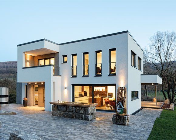 Nowoczesny dom z białymi ścianami, dużymi oknami i balkonem. Widoczne oświetlenie zewnętrzne, kamienne elementy dekoracyjne i brukowana ścieżka. Minimalistyczny styl.