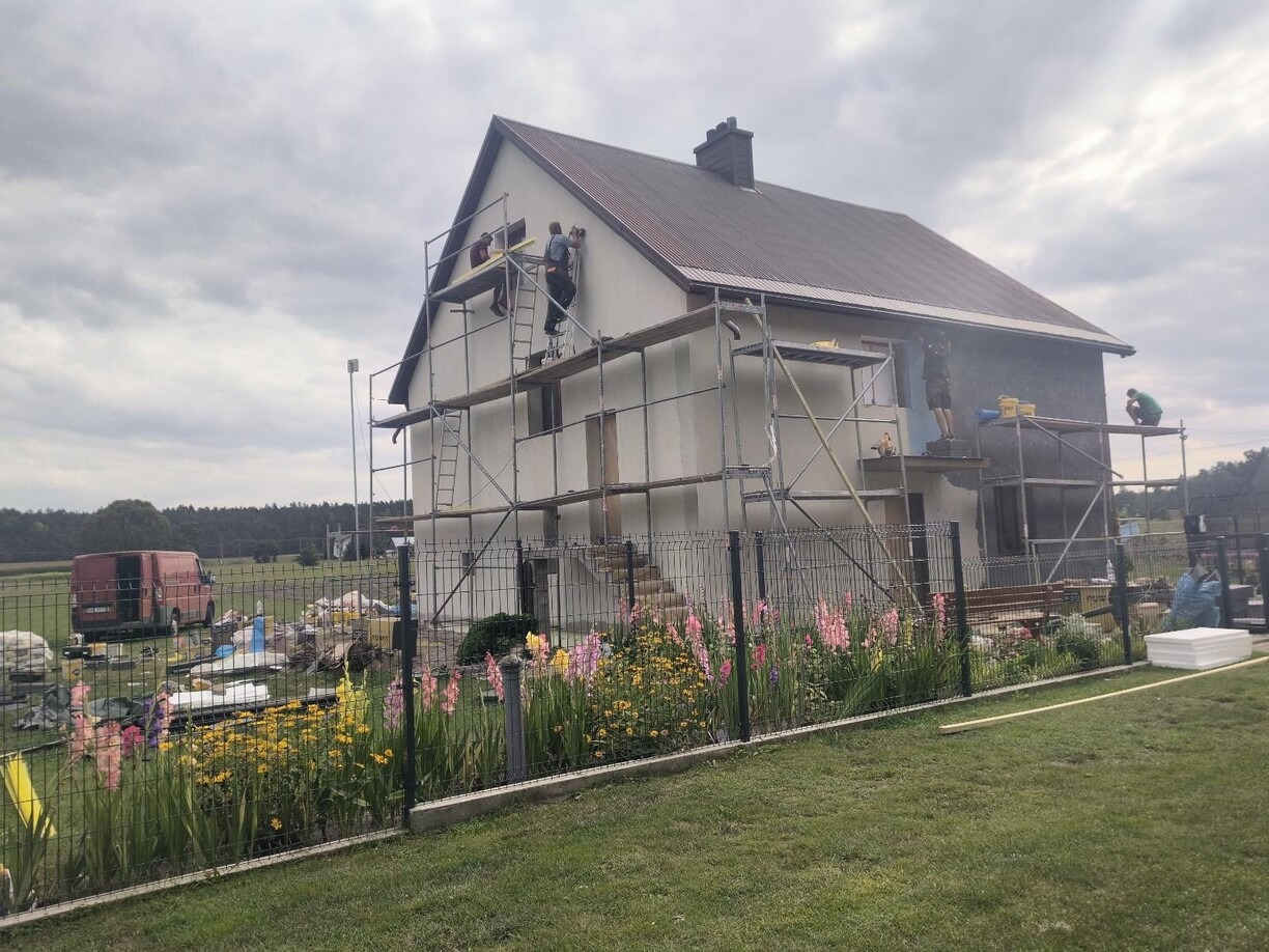 Dwukondygnacyjny dom w trakcie tynkowania elewacji; widoczne rusztowania, pracownicy i częściowo otynkowana ściana. W tle krajobraz i kwiaty w ogródku.